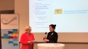 Zwei Frauen vor einer Präsentation und dem Roll-up des Wertebündnis Bayern, eine Dame spricht in ein Mikrofon, die andere hat Unterlagen in der Hand, sie lächelnt