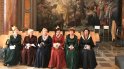 6 Damen in historischen Gewändern mit Kronen im Haar und kleinen Blumensträußen in der Hand sitzen da und lächeln