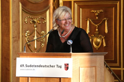 eine Person steht am Rednerpul und lacht, Im Hintergrund goldene Verzierungen, auf einem Schild am Rednerult steht 